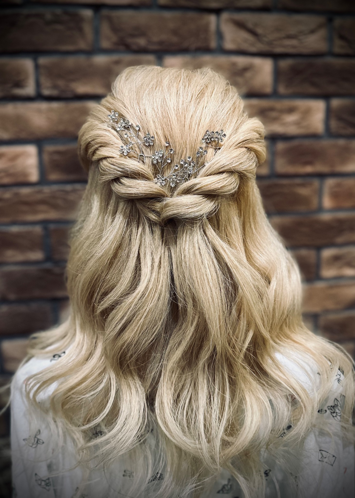 Upięcie blond włosów z warkoczem i ozdobnymi spinkami na tle ceglanej ściany, widok z tyłu.