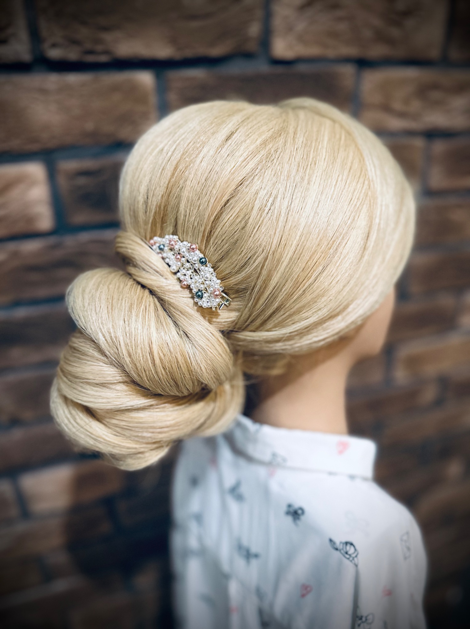 Blond włosy upięte w kok z ozdobną spinką z pereł i kamieni na tle ceglanej ściany, widok z tyłu.