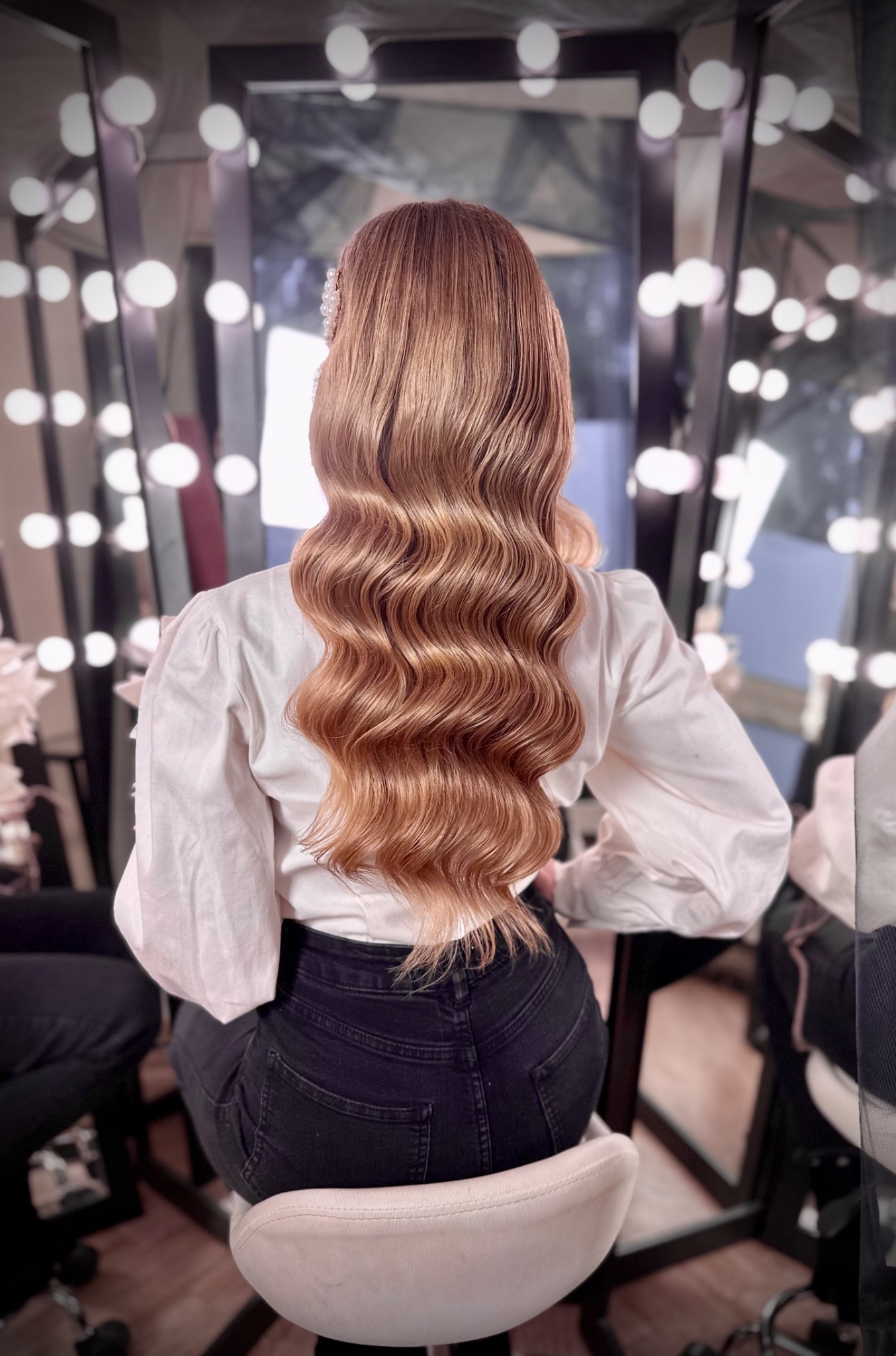 Widok z tyłu na długie, falowane, miedziano-blond włosy z ozdobą z pereł, biała bluzka z bufiastymi rękawami, czarne dżinsy, w tle lustro z oświetleniem