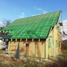 Budowa domu w Żninie: widok na częściowo pokryty zieloną dachówką dach z widocznymi elementami konstrukcyjnymi i rusztowaniem, etap prac w słoneczny dzień.