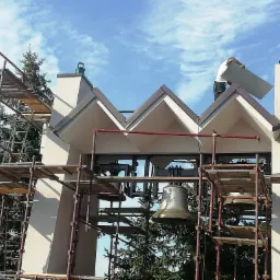 Pracownik na rusztowaniu montuje element na dachu wieży kościelnej z dzwonami, na tle błękitnego nieba.