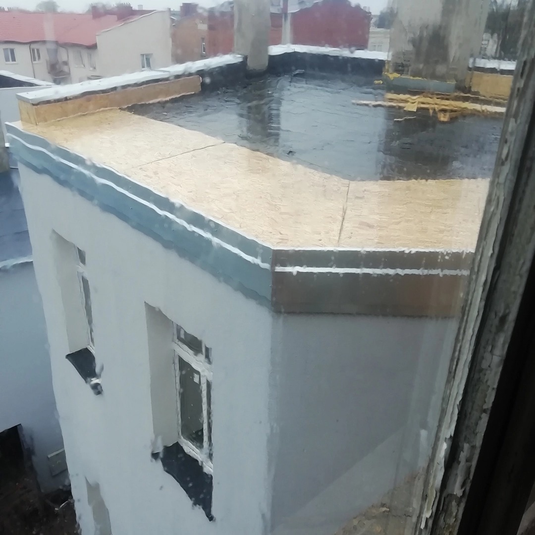 Płaski dach budynku po deszczu, widoczne nowe obłożenie z płyt OSB i obróbki blacharskie, z perspektywy okna wewnątrz budynku.