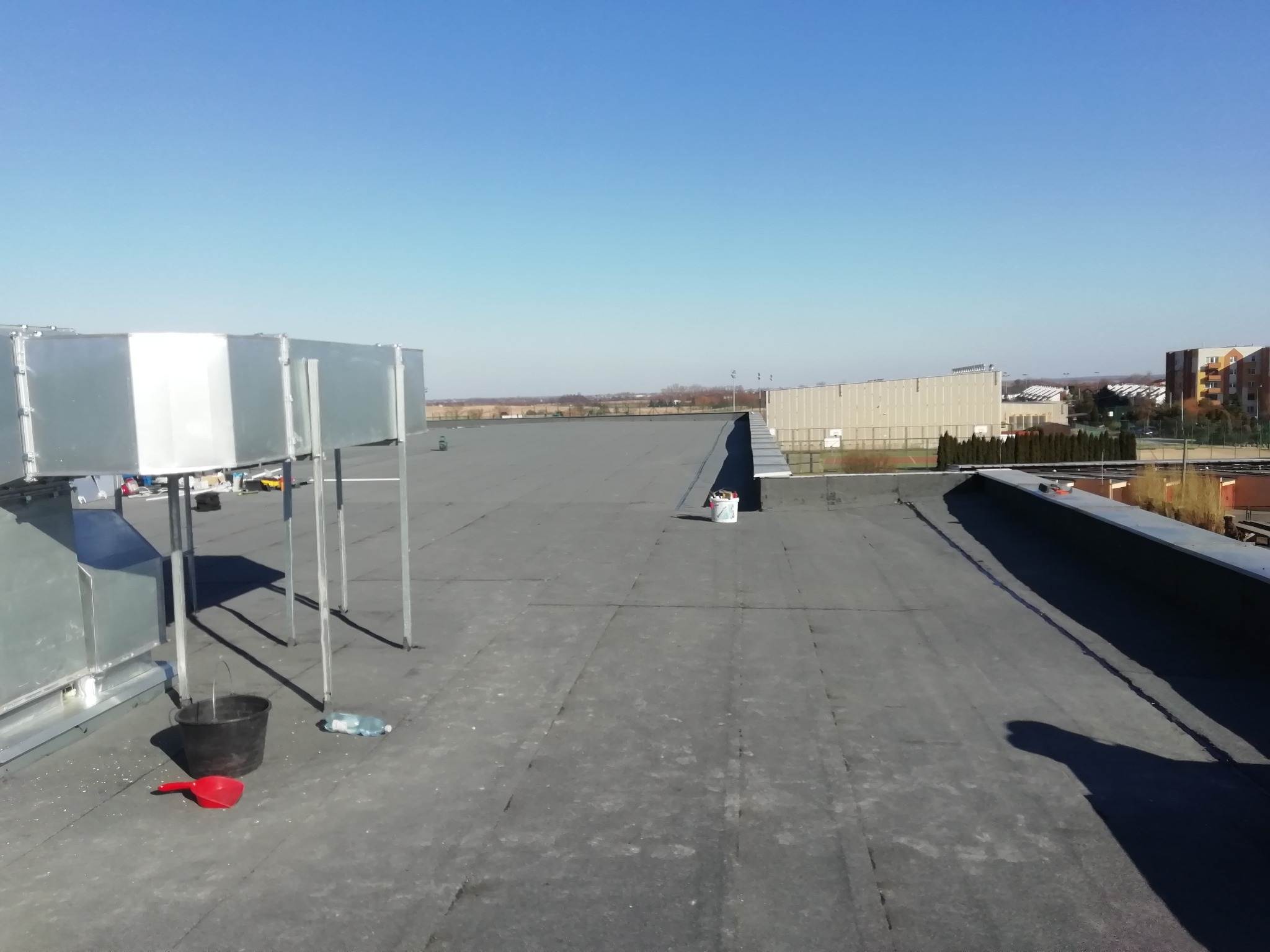 Rozległy płaski dach pokryty papą, z widocznymi elementami wentylacji, wiadrem, czerwoną szpachelką i butelką wody, w tle budynki i błękitne niebo.