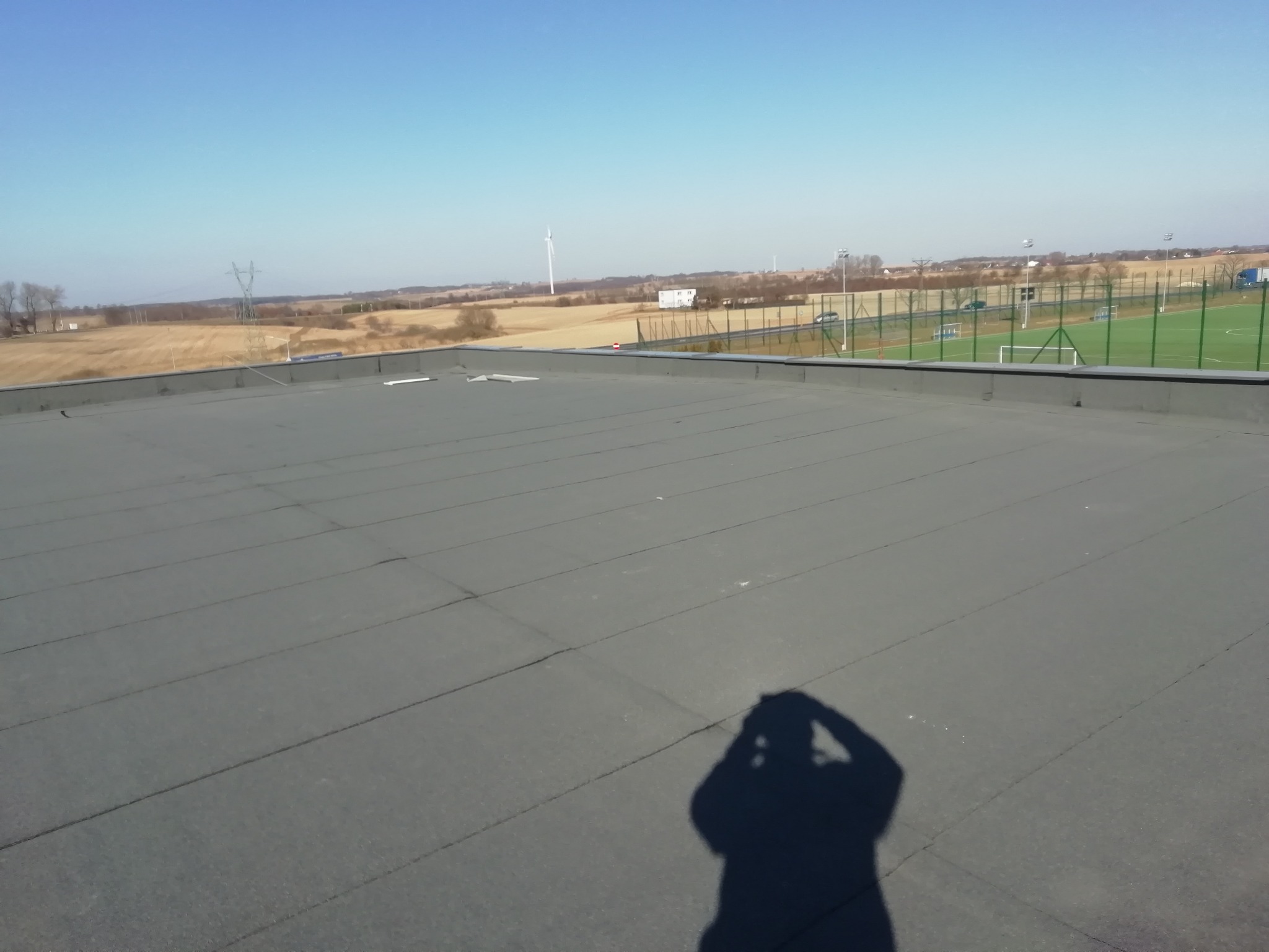 Płaski dach pokryty papą termozgrzewalną z widocznym cieniem fotografa, w tle krajobraz z polami, turbiną wiatrową i boiskiem sportowym.