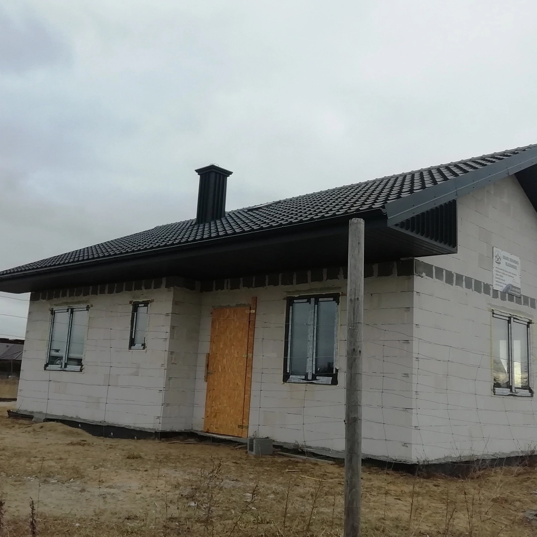 Niedokończony dom z ciemnym dachem z dachówki i kominem, widok z boku na tle pochmurnego nieba.