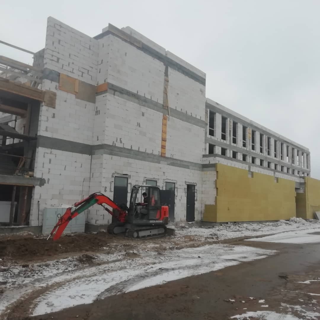 Budowa budynku z białych bloczków, widoczna mała koparka na tle ocieplonej ściany i fragment dachu w trakcie montażu.