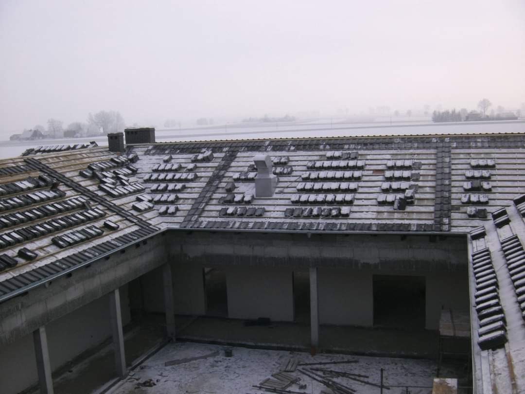 Niedokończone krycie dachu budynku gospodarczego z wewnętrznym dziedzińcem, częściowo pokryte śniegiem, widok z góry na ułożone dachówki i kominy na tle zimowego krajobrazu.