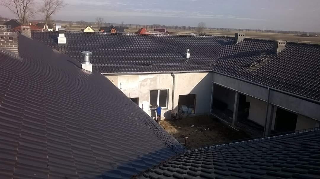 Widok z góry na nowo położony, ciemny dach z blachodachówki na budynku w trakcie wykończenia elewacji, z widocznymi kominami i pracownikami na dziedzińcu.