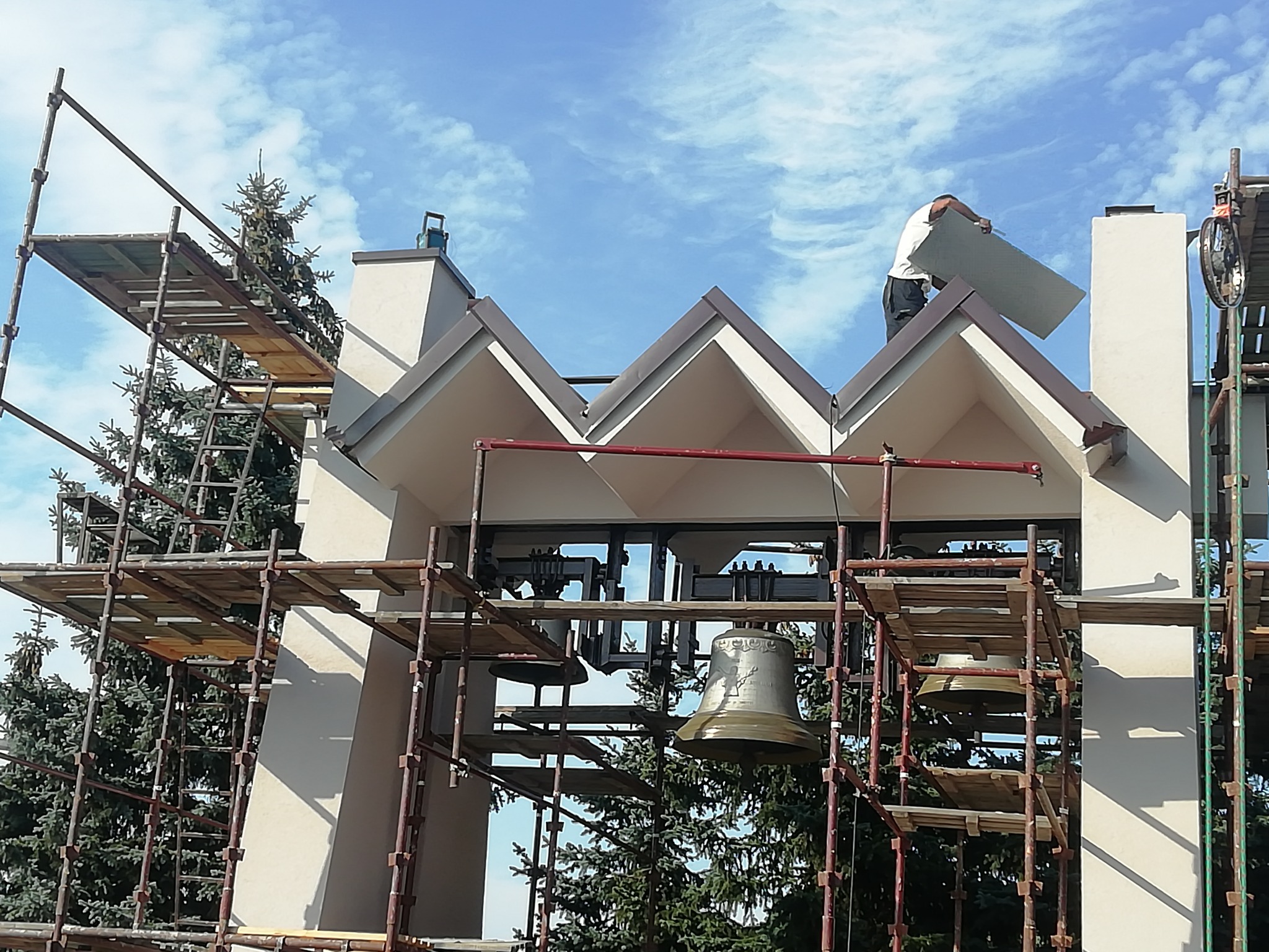 Pracownik na rusztowaniu montuje element na dachu wieży kościelnej z dzwonami, na tle błękitnego nieba.