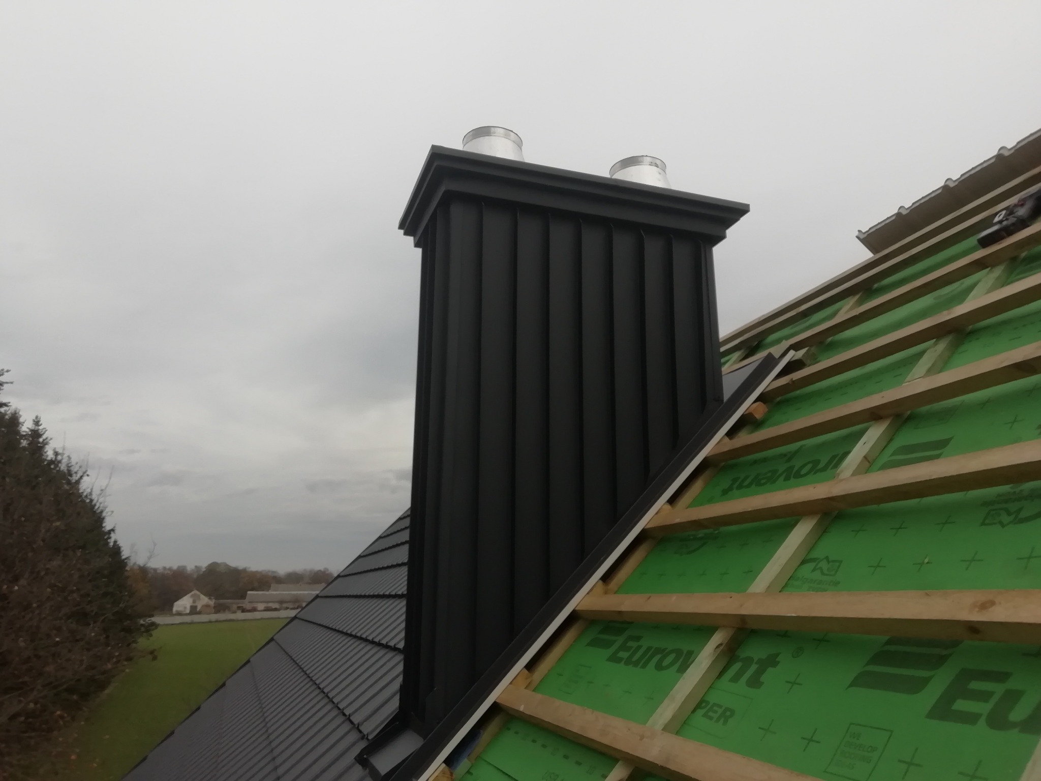 Ciemnoszary komin z dwoma metalowymi wylotami na tle pochmurnego nieba, widok z dachu w trakcie prac dekarskich, z widoczną konstrukcją drewnianą i membraną dachową.