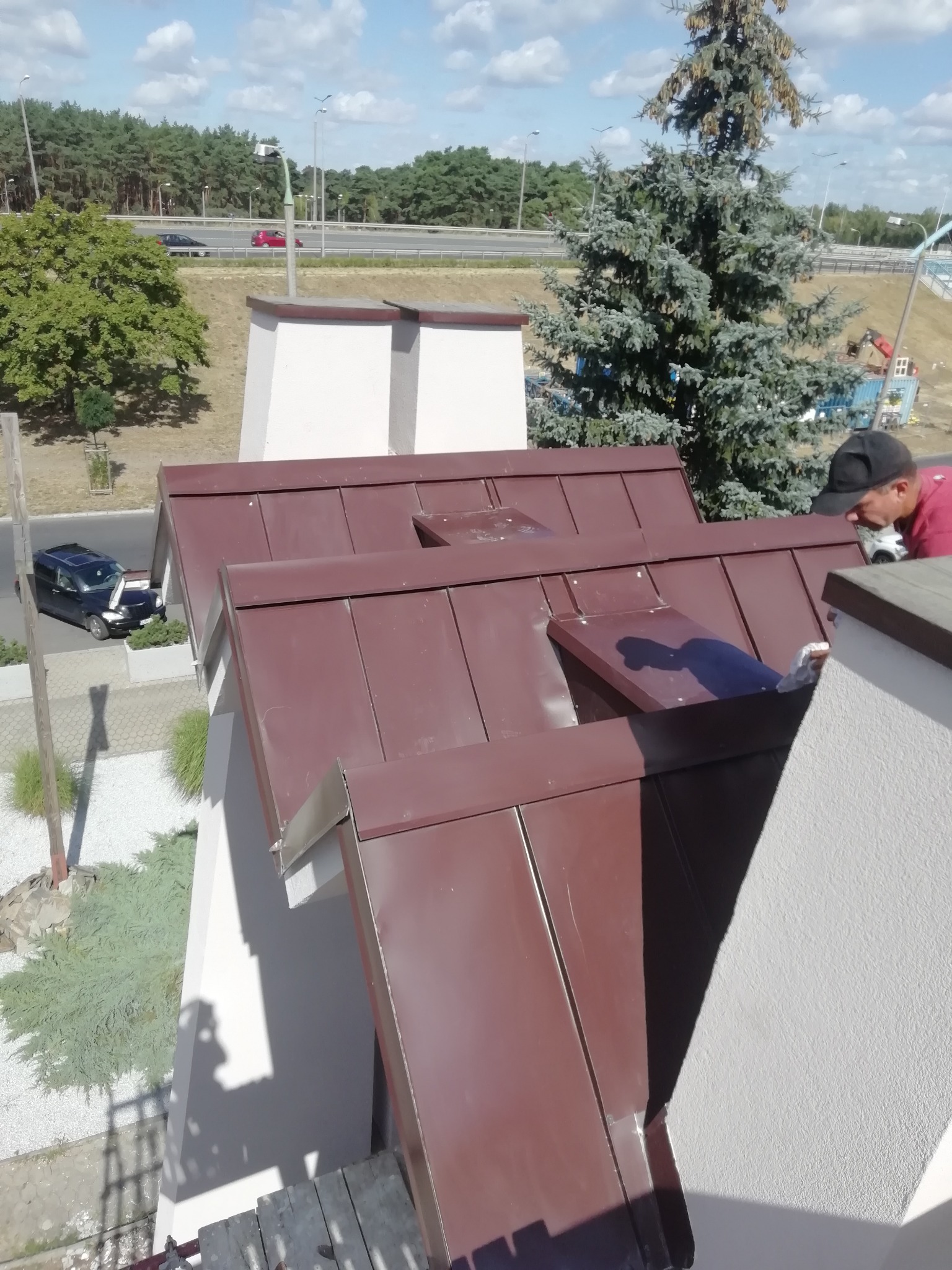 Widok z góry na fragment dachu z brązowej blachy trapezowej, z widocznymi kominami i oknami dachowymi, na tle ruchliwej drogi szybkiego ruchu i zieleni, z dekarzem w czapce przy pracy.