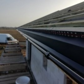 Czarna, metalowa rynna zamontowana na budynku w trakcie budowy, widoczne krokwie dachu i pustaki. W tle krajobraz z drzewami i samochodem dostawczym.