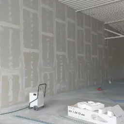 Montaż płyt karton-gips w hali w Prudniku. Widoczne łączenia płyt i szpachlowanie, sprzęt budowlany i pracownik w oddali.