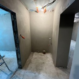 Eagle Eye - Szara łazienka w trakcie remontu: świeżo położone gładzie na ścianach, widoczne instalacje elektryczne pod sufitem zabezpieczonym folią. Otwór drzwiowy z ościeżnicą.