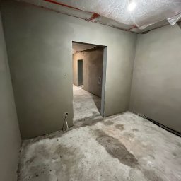 Eagle Eye - Wnętrze w trakcie prac wykończeniowych: świeżo otynkowane ściany w szarym odcieniu, zabezpieczona folią sufitowa z żarówką, widoczne kable i surowa podłoga.