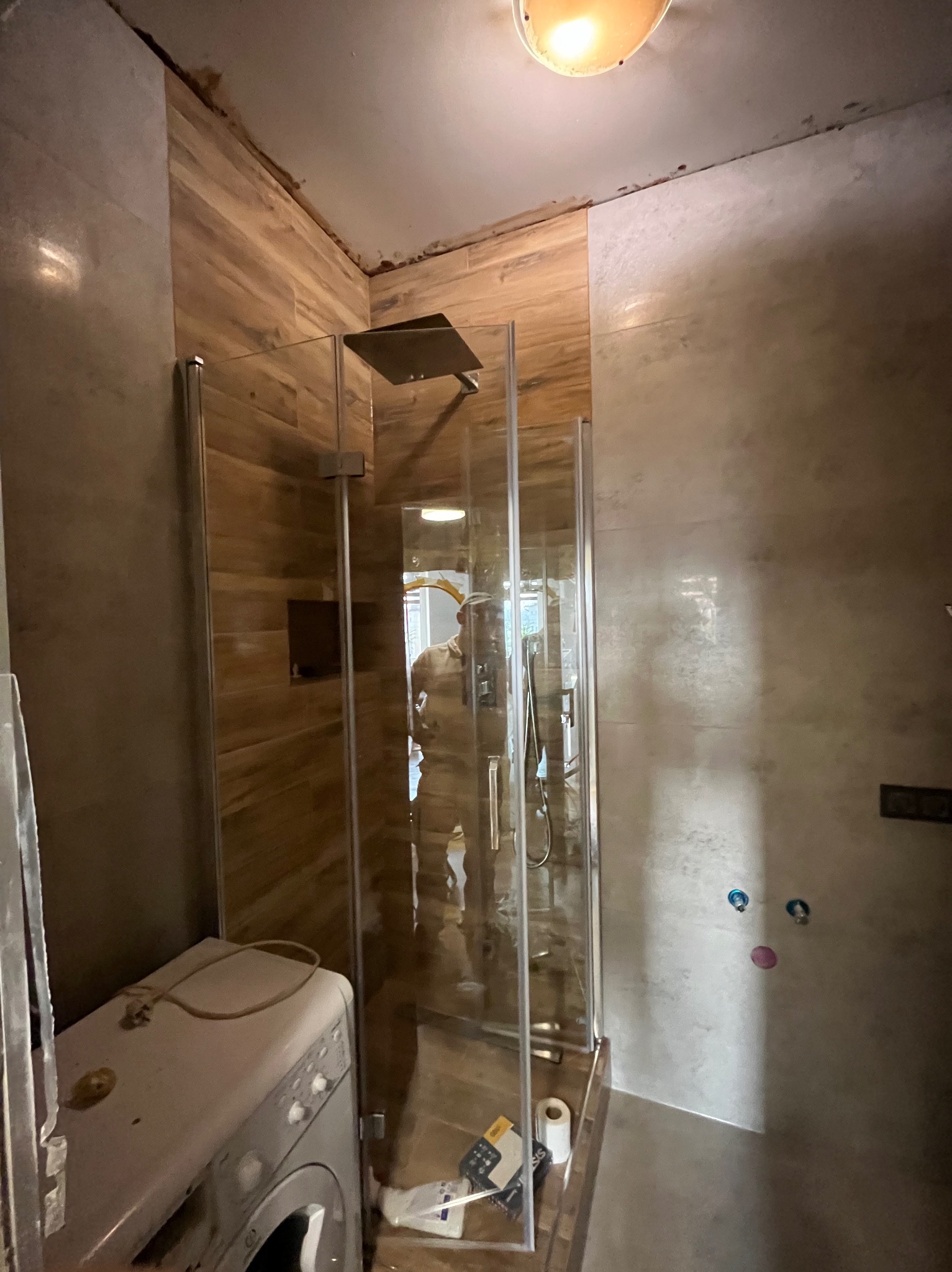 Wnętrze łazienki w trakcie remontu. Widoczna kabina prysznicowa z deszczownicą, pralka i częściowo wykończone ściany z płytek imitujących drewno i kamień.