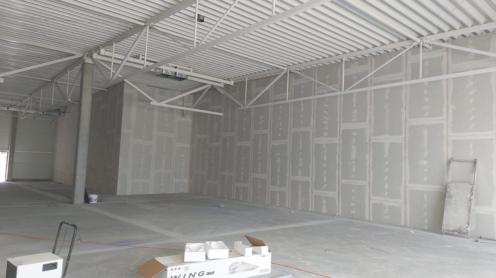 Montaż płyt kartonowo-gipsowych na ścianach wewnątrz hali, widoczne łączenia i szpachlowanie. Stalowa konstrukcja dachu, surowy beton na podłodze, przygotowania do dalszych prac.