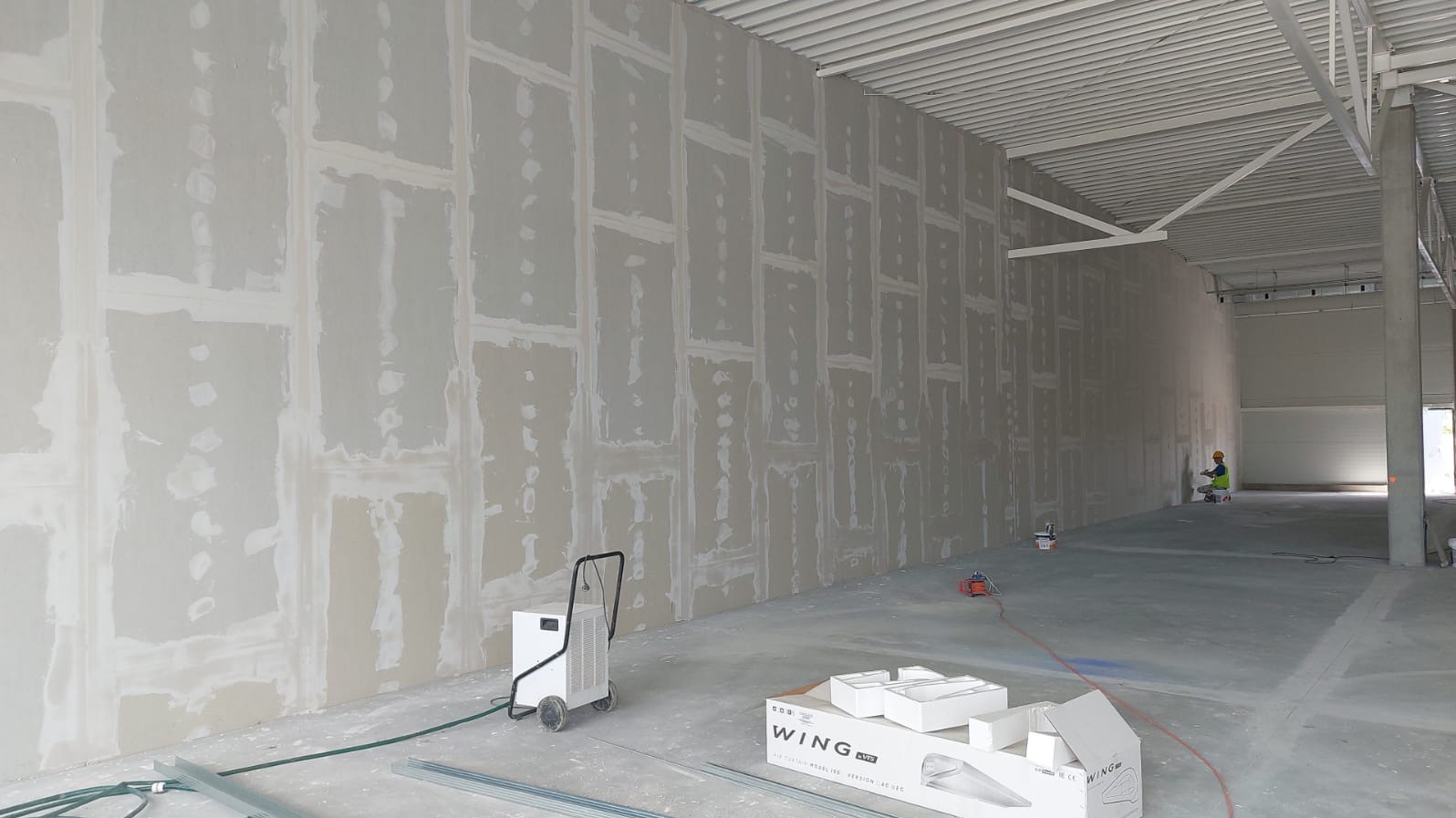 Montaż płyt karton-gips w hali w Prudniku. Widoczne łączenia płyt i szpachlowanie, sprzęt budowlany i pracownik w oddali.