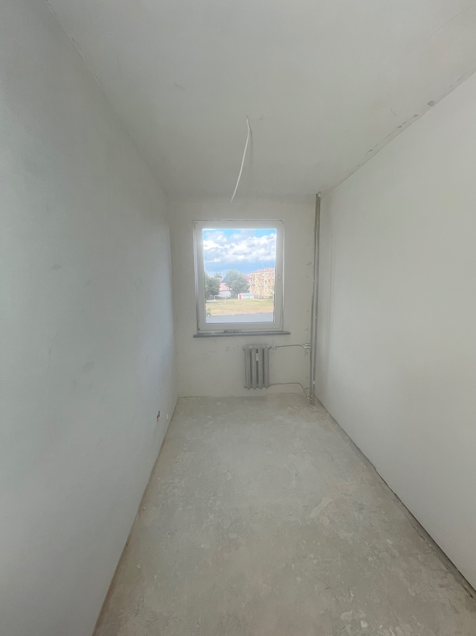 Pusty pokój w trakcie remontu: białe ściany, surowy beton na podłodze, grzejnik pod oknem z widokiem na budynek. Widoczne ślady po gładzi.