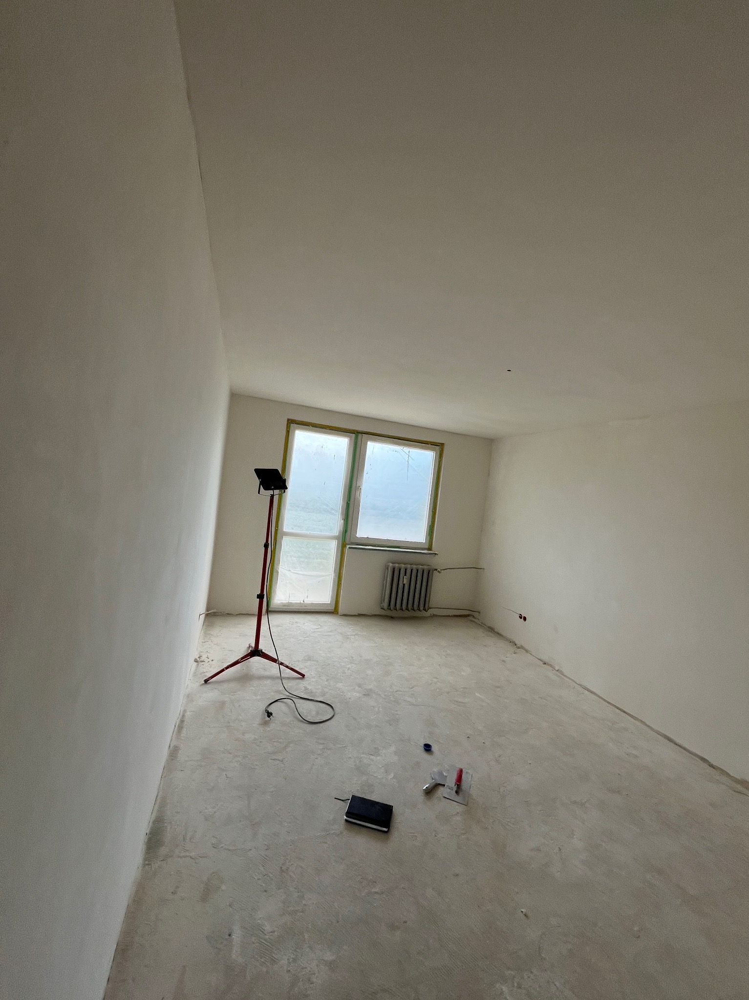 Pokój po gładziowaniu, przygotowany do malowania. Widoczne narzędzia, lampa oświetleniowa i zabezpieczone okno folią. Ściany gładkie, jasne, gotowe na kolejny etap.