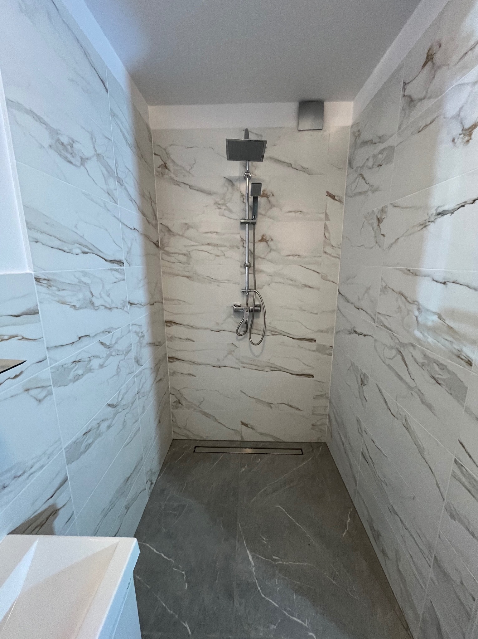 Nowoczesna kabina prysznicowa z marmurowymi płytkami ściennymi i szarą podłogą. Minimalistyczny design z odpływem liniowym i deszczownicą.