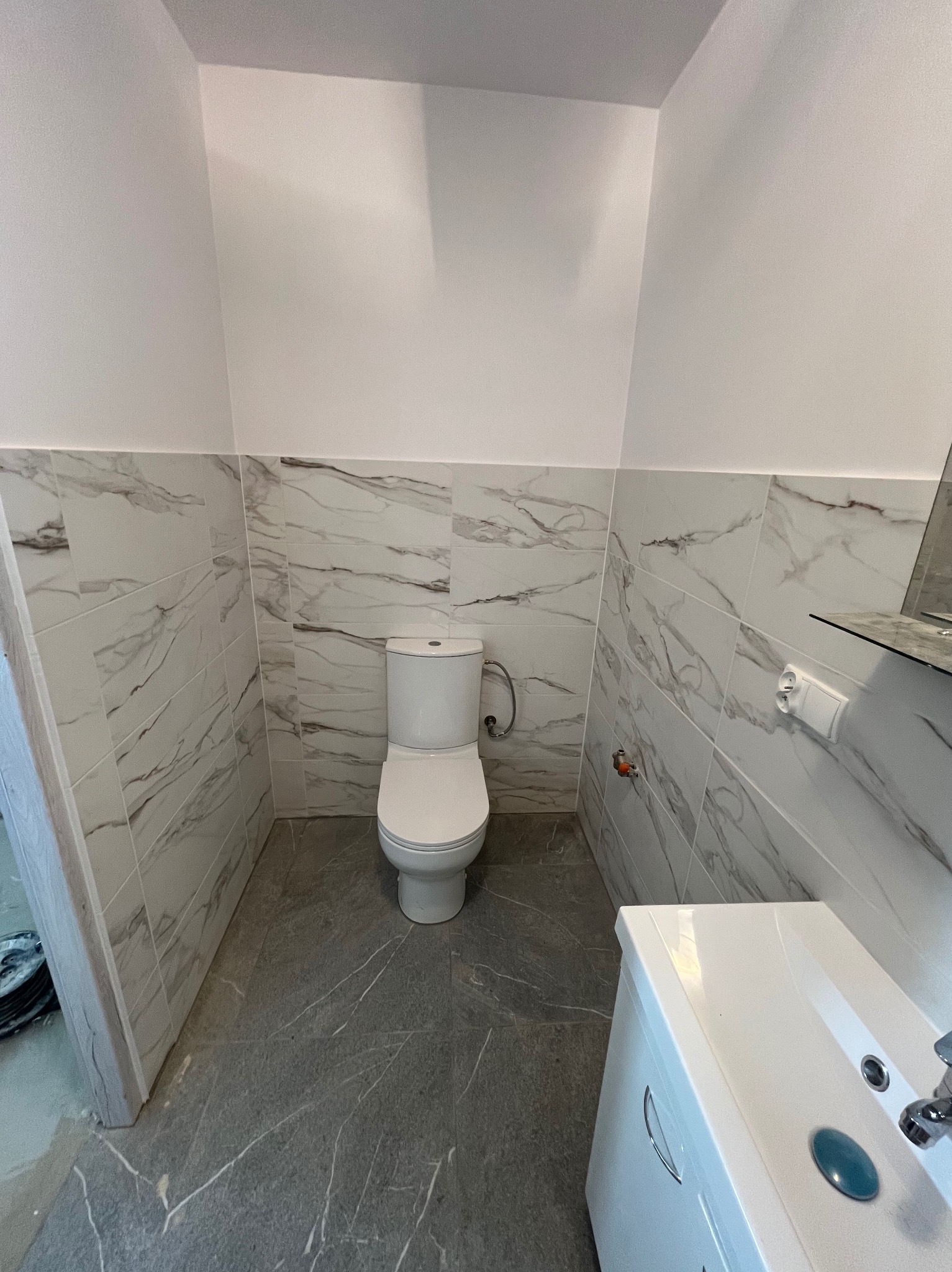 Wykończona łazienka z marmurowymi płytkami na ścianach i podłodze. Widoczna toaleta, umywalka z szafką i gniazdko elektryczne. Minimalistyczny design.