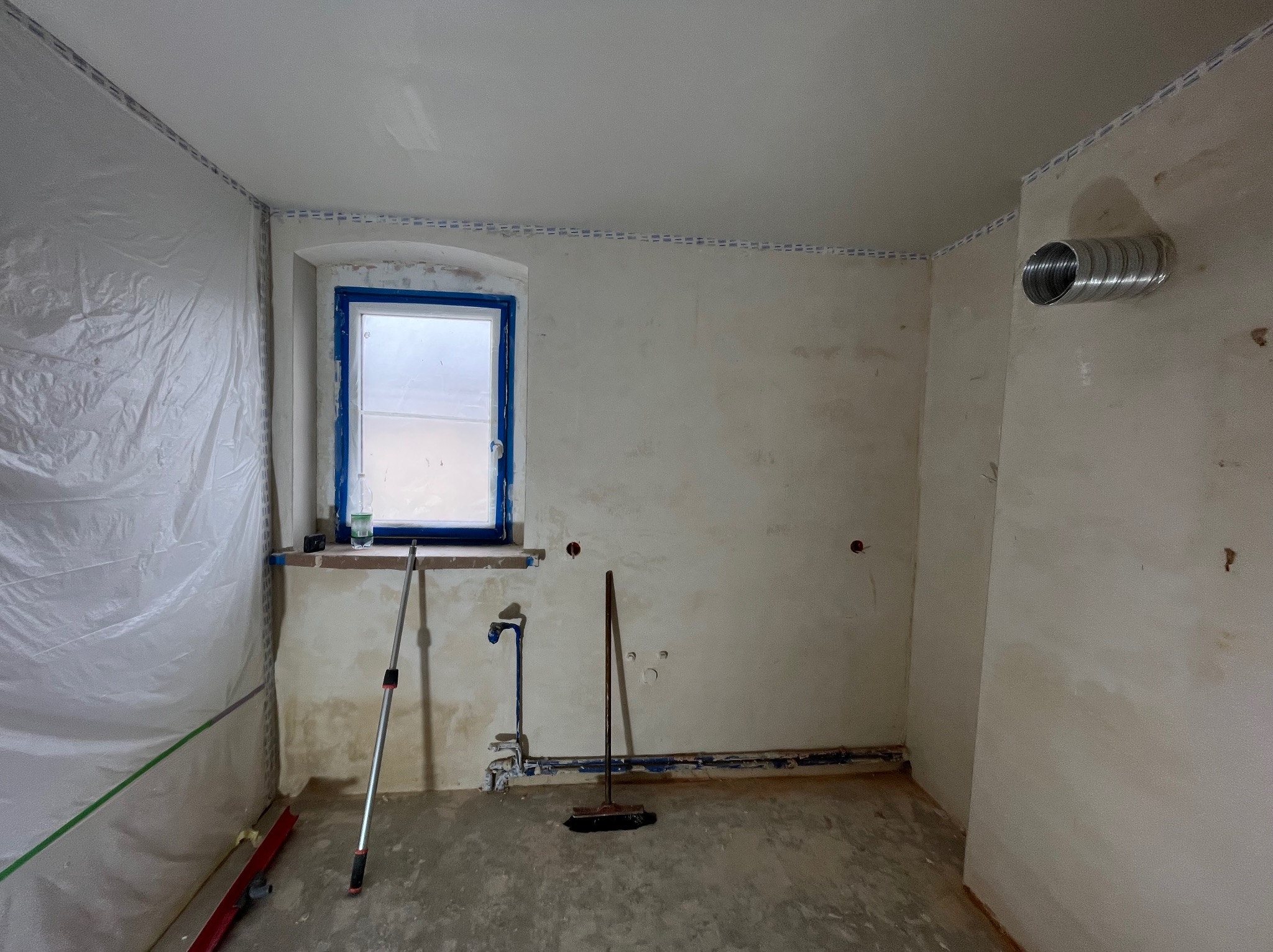 Wnętrze w trakcie remontu: okno z niebieską ramą, ściany przygotowane do malowania, zabezpieczone folią, widoczne narzędzia i instalacje.