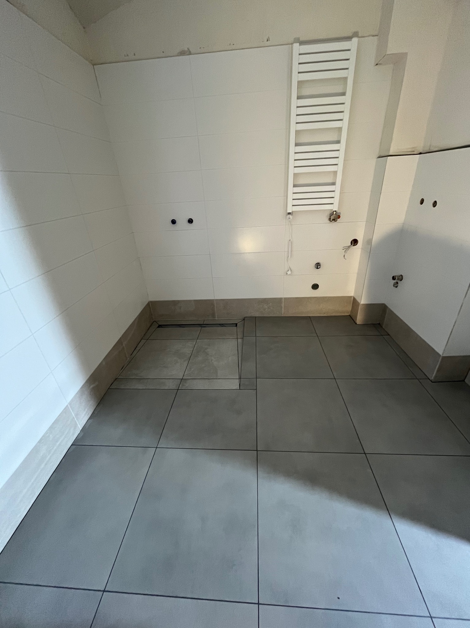 Wykończona łazienka z szarymi płytkami podłogowymi i białymi płytkami ściennymi. Widoczny grzejnik drabinkowy i przygotowane punkty instalacyjne.