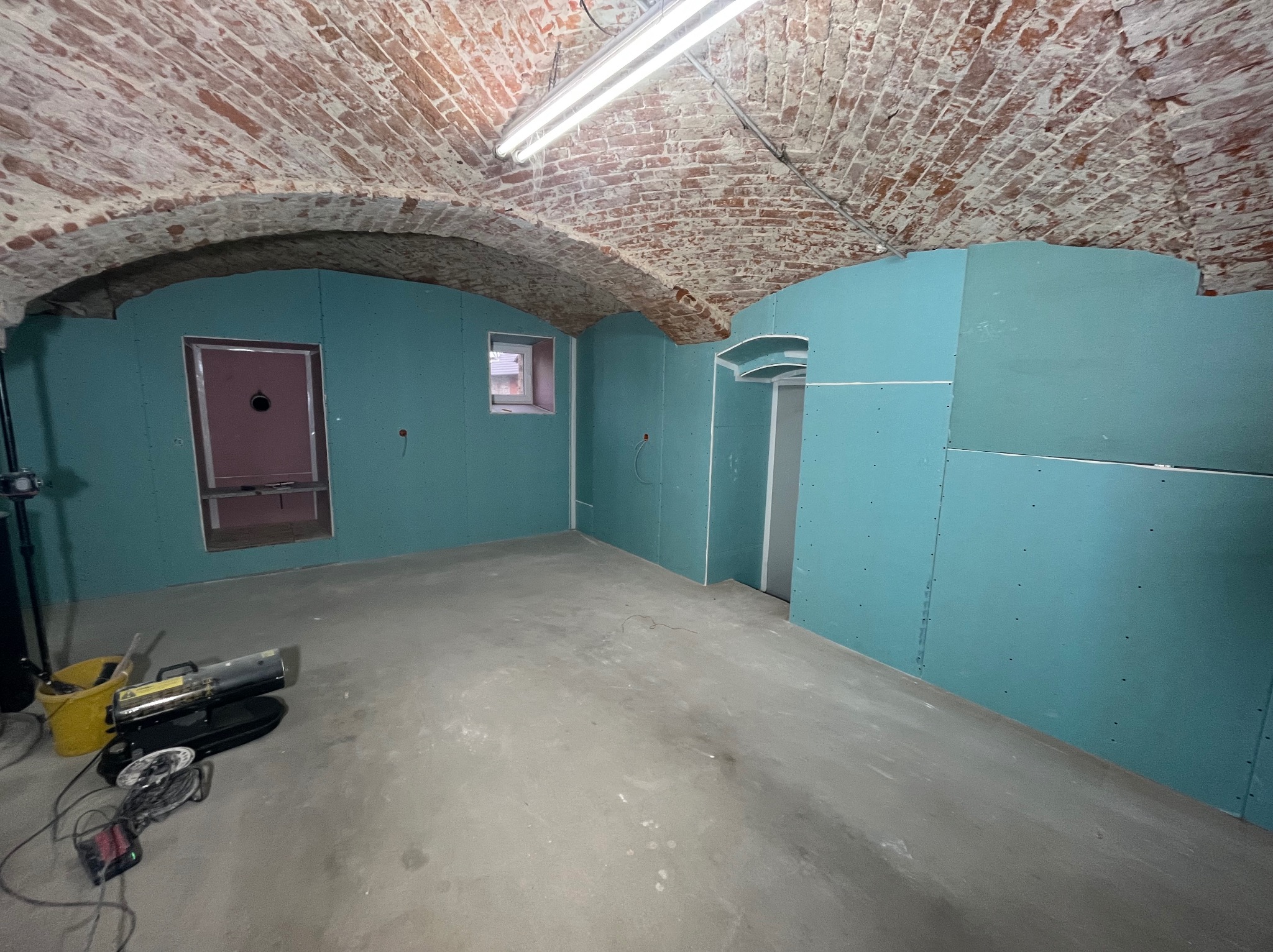 Montaż płyt G-K na ścianach w Prudniku. Widoczny sufit z cegły, narzędzia i przygotowane otwory na okna i drzwi. Etap prac wykończeniowych w pomieszczeniu.