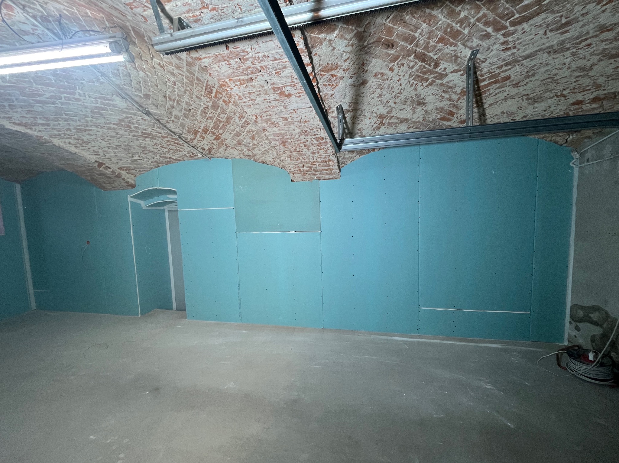 Montaż płyt gipsowo-kartonowych na ścianach w pomieszczeniu z ceglanym sklepieniem łukowym. Widoczne profile montażowe i instalacja elektryczna.