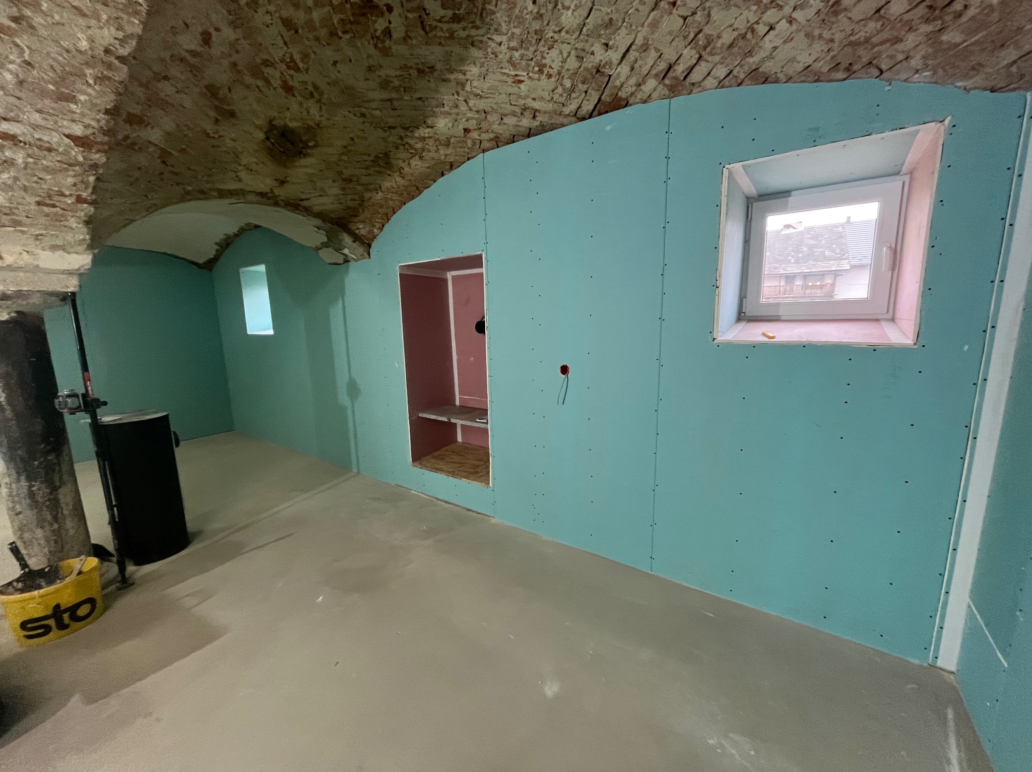 Niebieskie płyty gipsowo-kartonowe zamontowane na ścianach z oknem i wnęką w pomieszczeniu z ceglanym sklepieniem. Widoczne narzędzia i wiadro.