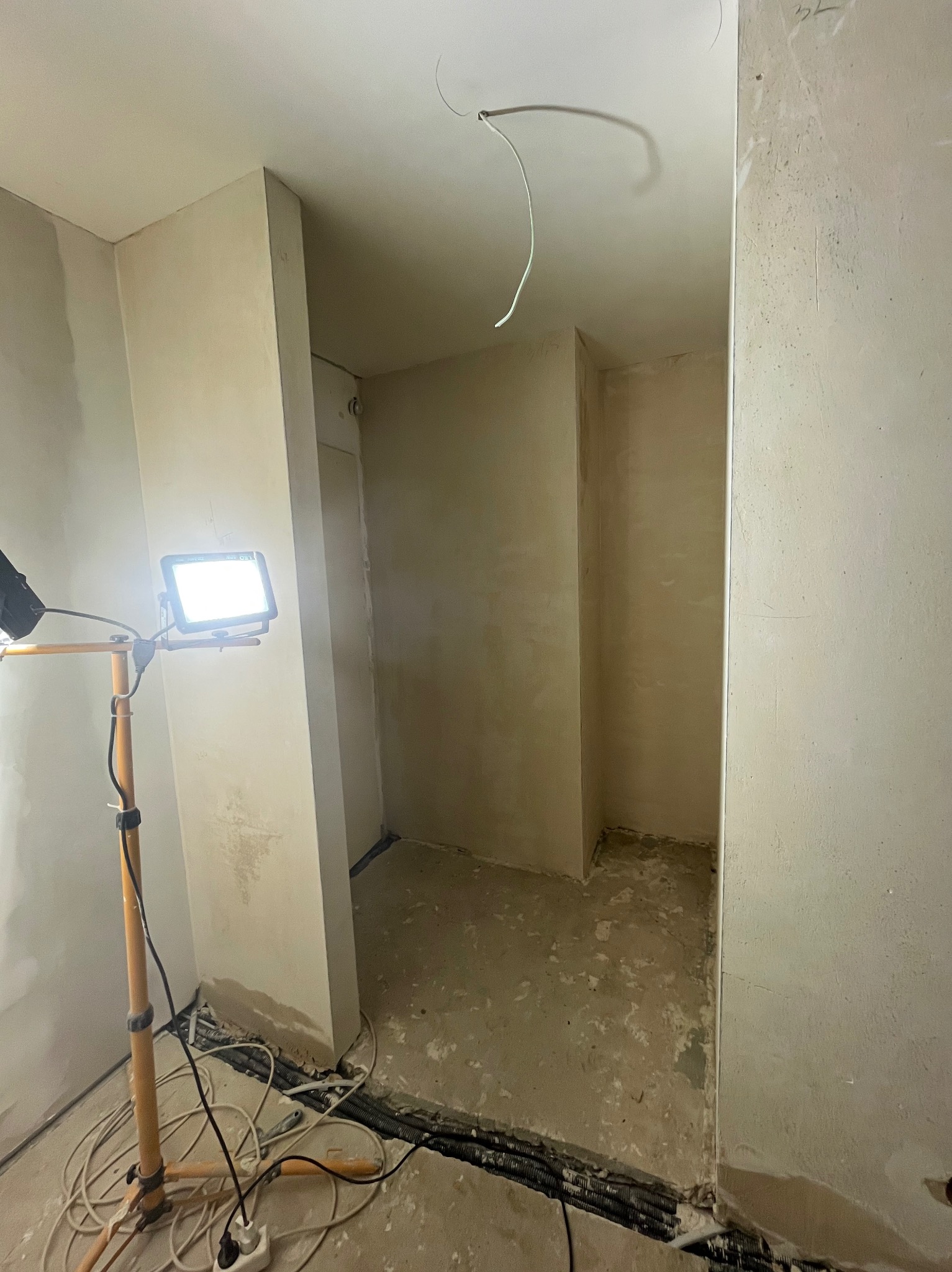 Wnętrze w trakcie prac wykończeniowych: ściany po tynkowaniu, widoczny przewód elektryczny i lampa budowlana na statywie. Podłoga surowa, przygotowana do dalszych prac.