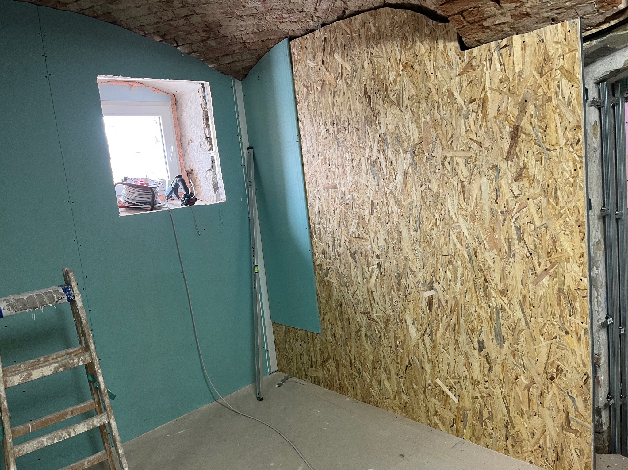 Wnętrze w trakcie prac wykończeniowych: ściany z płyt gipsowo-kartonowych w odcieniu turkusu, fragment ściany z płyty OSB, drabina malarska i okno z narzędziami.