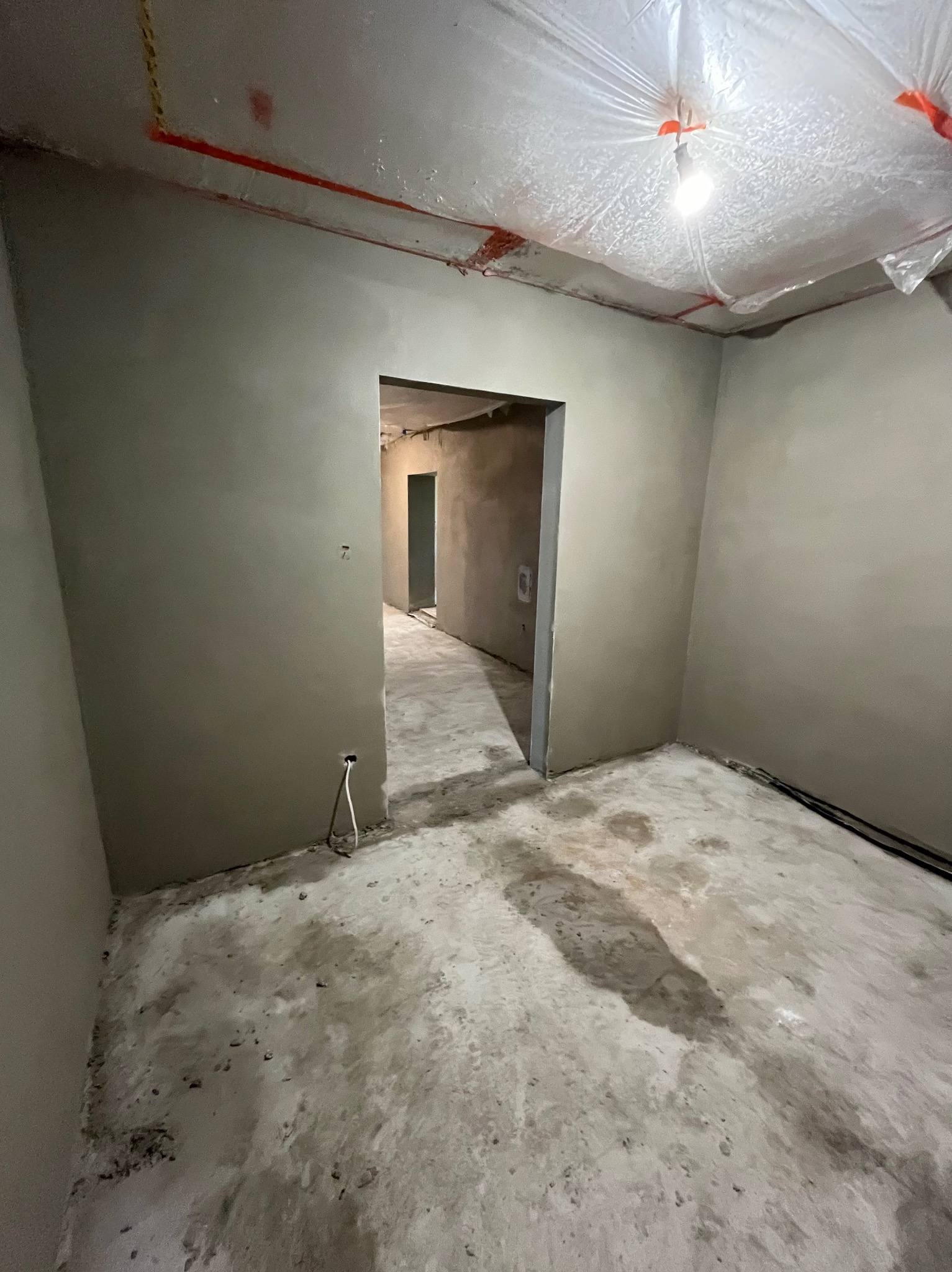 Wnętrze w trakcie prac wykończeniowych: świeżo otynkowane ściany w szarym odcieniu, zabezpieczona folią sufitowa z żarówką, widoczne kable i surowa podłoga.