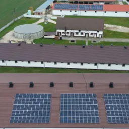 Widok z góry na budynki gospodarcze z panelami słonecznymi na brązowym dachu, otoczone zielonym polem i innymi budynkami.