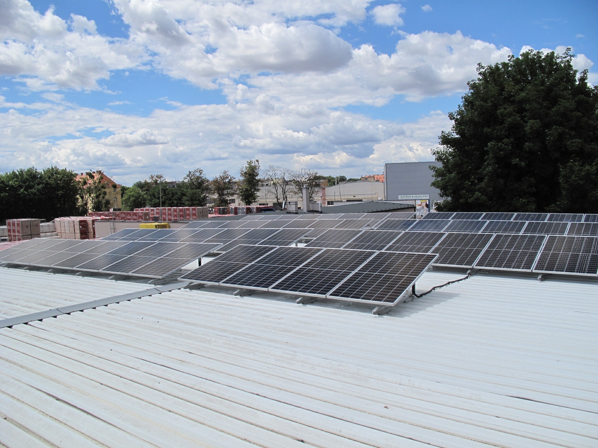 Panele fotowoltaiczne zamontowane na białym, blaszanym dachu budynku, widoczne kable, w tle drzewa i budynki pod błękitnym niebem z chmurami.