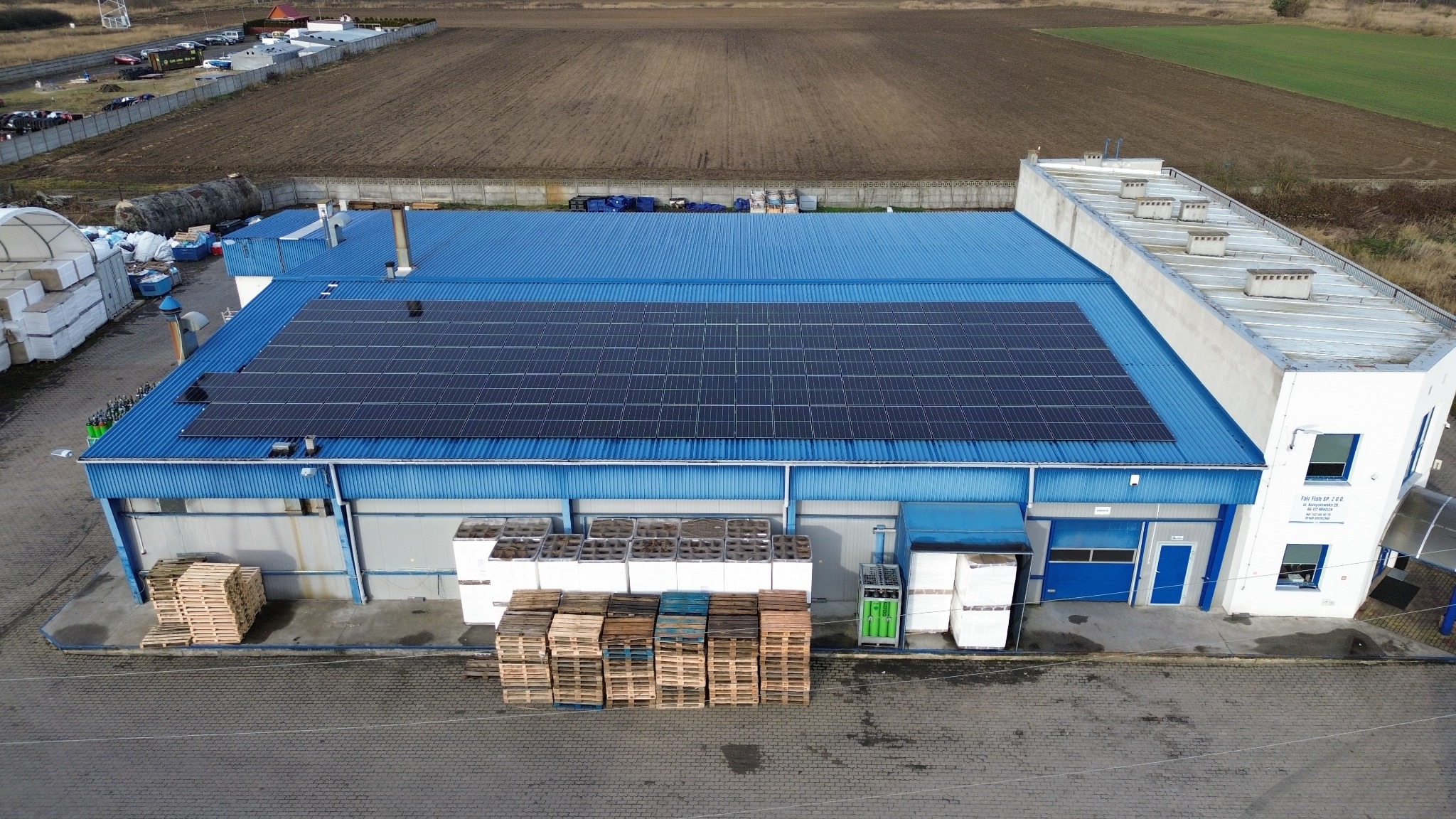 Widok z góry na budynek przemysłowy z niebieskim dachem, na którym zamontowane są panele słoneczne. Na placu przed budynkiem składowane palety i białe kontenery.