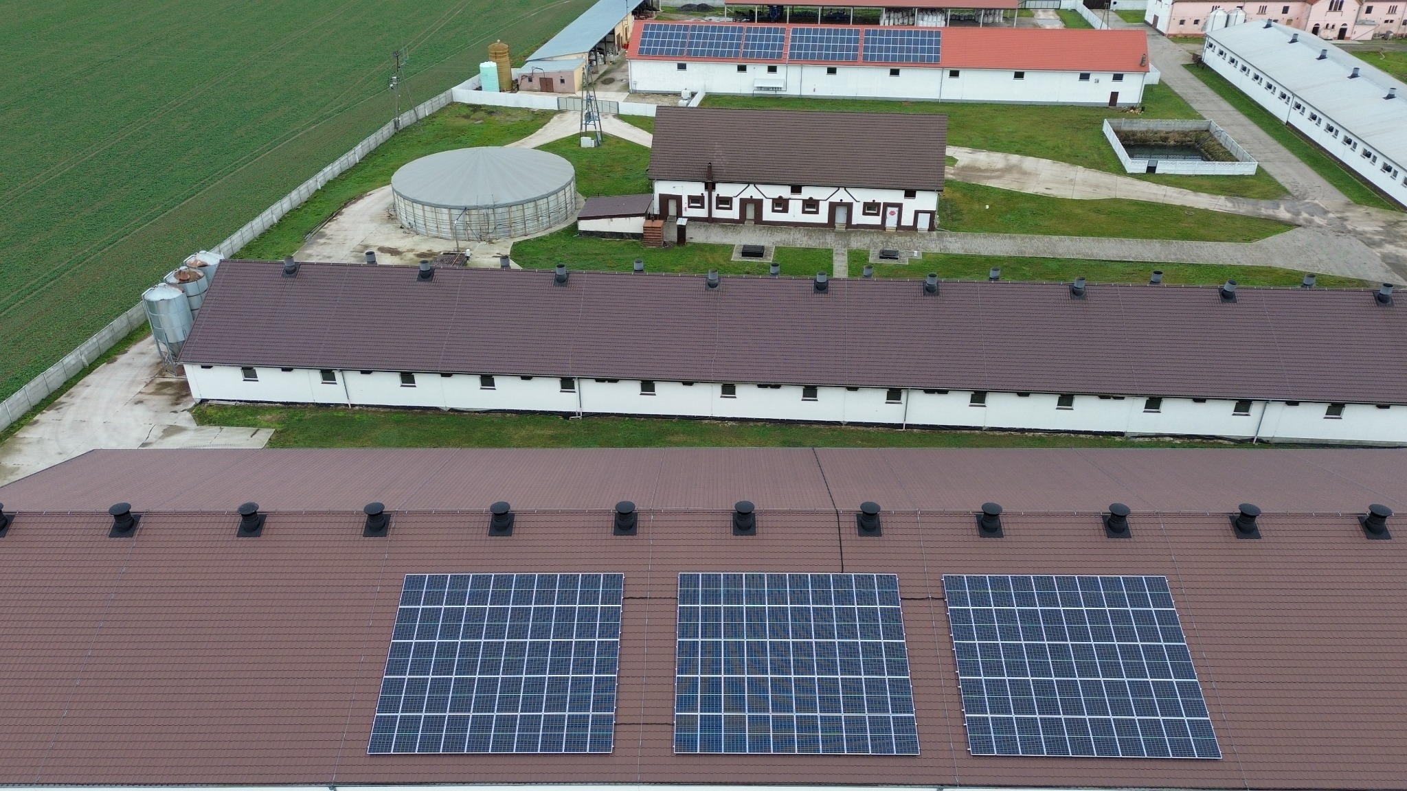 Widok z góry na budynki gospodarcze z panelami słonecznymi na brązowym dachu, otoczone zielonym polem i innymi budynkami.