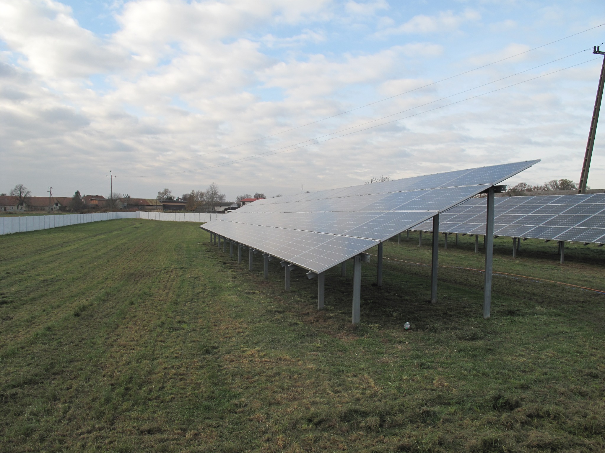 Rząd paneli słonecznych na metalowych podporach na trawiastym polu, z widocznymi liniami energetycznymi i słupem w tle, pod pochmurnym niebem.