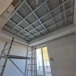 Metalowa konstrukcja sufitowa z widocznymi przewodami elektrycznymi, zamontowana w surowym wnętrzu z drabiną i oknem balkonowym.