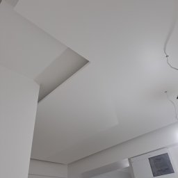 Yevhenii Tolmachov - Biały sufit podwieszany z wnęką oświetleniową i wnęką na instalacje, widoczna skrzynka elektryczna na ścianie.