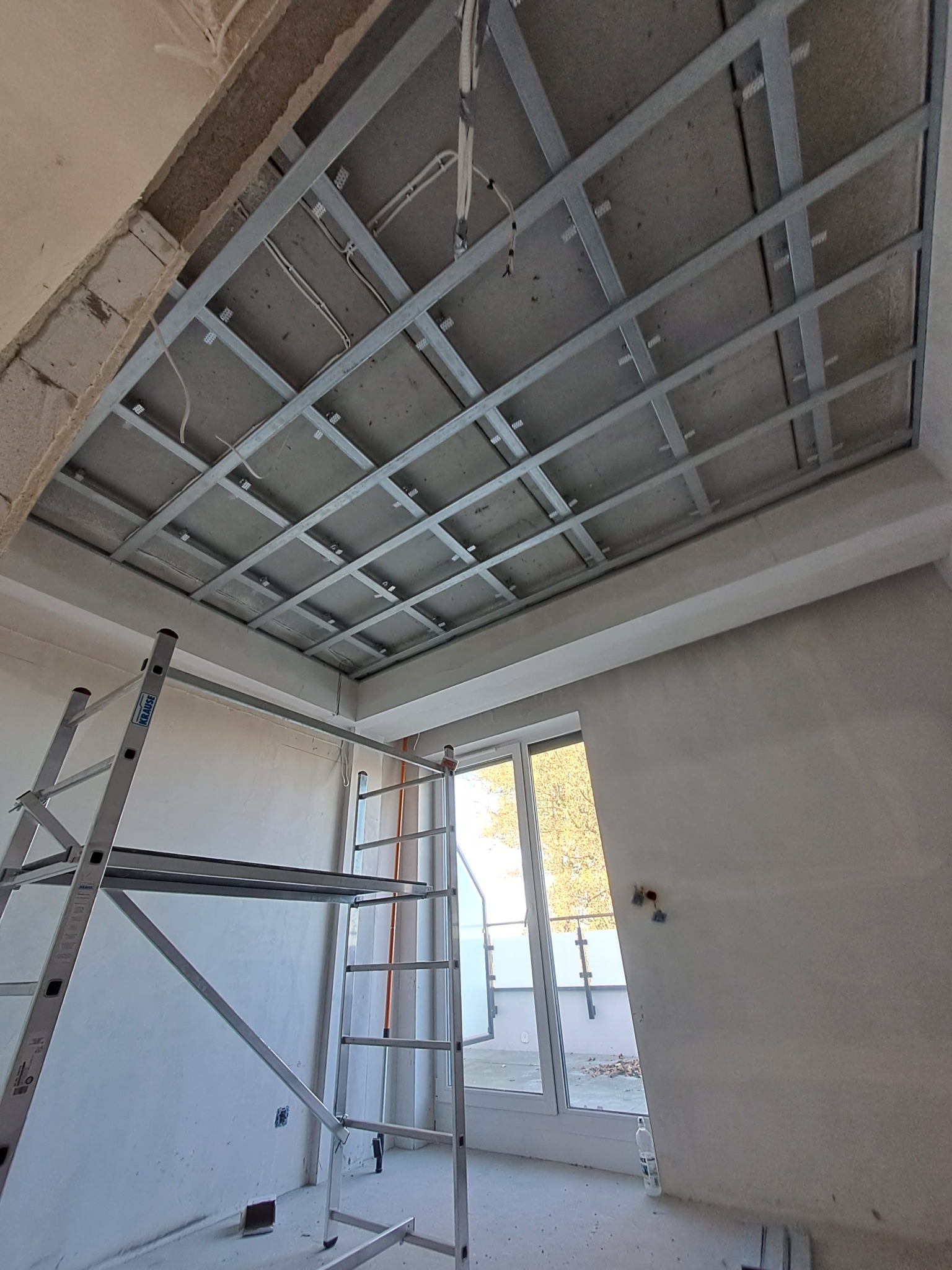 Metalowa konstrukcja sufitowa z widocznymi przewodami elektrycznymi, zamontowana w surowym wnętrzu z drabiną i oknem balkonowym.