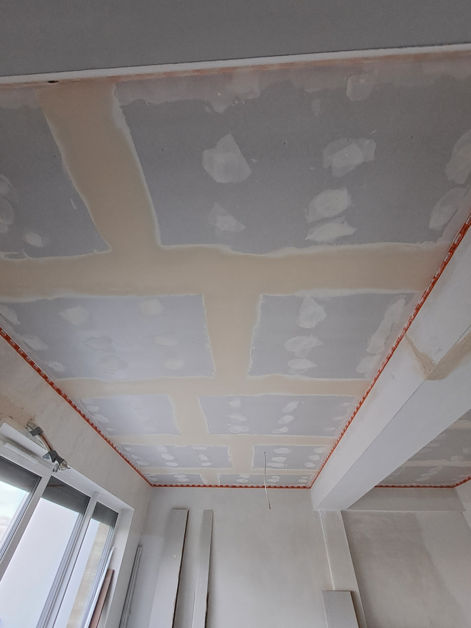 Sufit podwieszany z płyt kartonowo-gipsowych w trakcie szpachlowania łączeń, widoczne pomarańczowe profile montażowe i niedoskonałości w szpachlowaniu, okno z lewej strony.