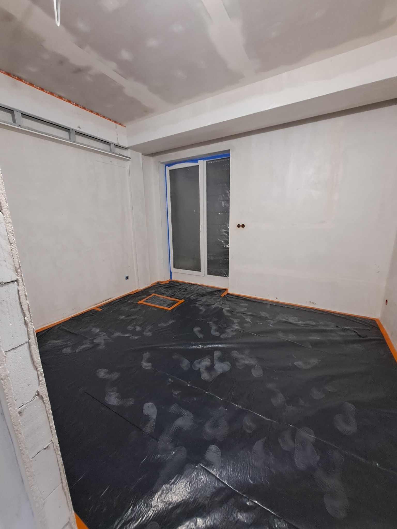 Przygotowanie do prac wykończeniowych we Wrocławiu: Pokryta folią podłoga w pomieszczeniu ze świeżo wyszpachlowanymi ścianami i sufitem, oklejone okno, widoczne profile pod płytę karton-gips.