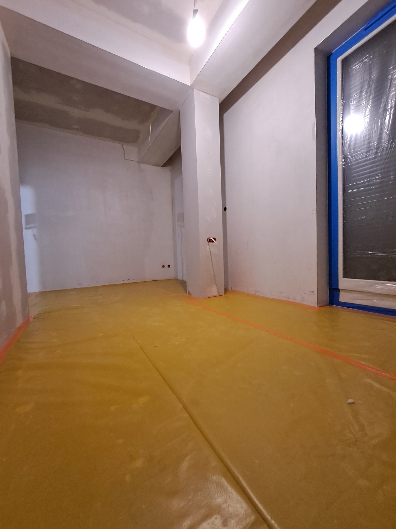 Pokój w trakcie przygotowań do prac wykończeniowych: zabezpieczona folią podłoga, ściany po wstępnym wyrównaniu, okno z niebieską taśmą malarską, widoczne puszki elektryczne.