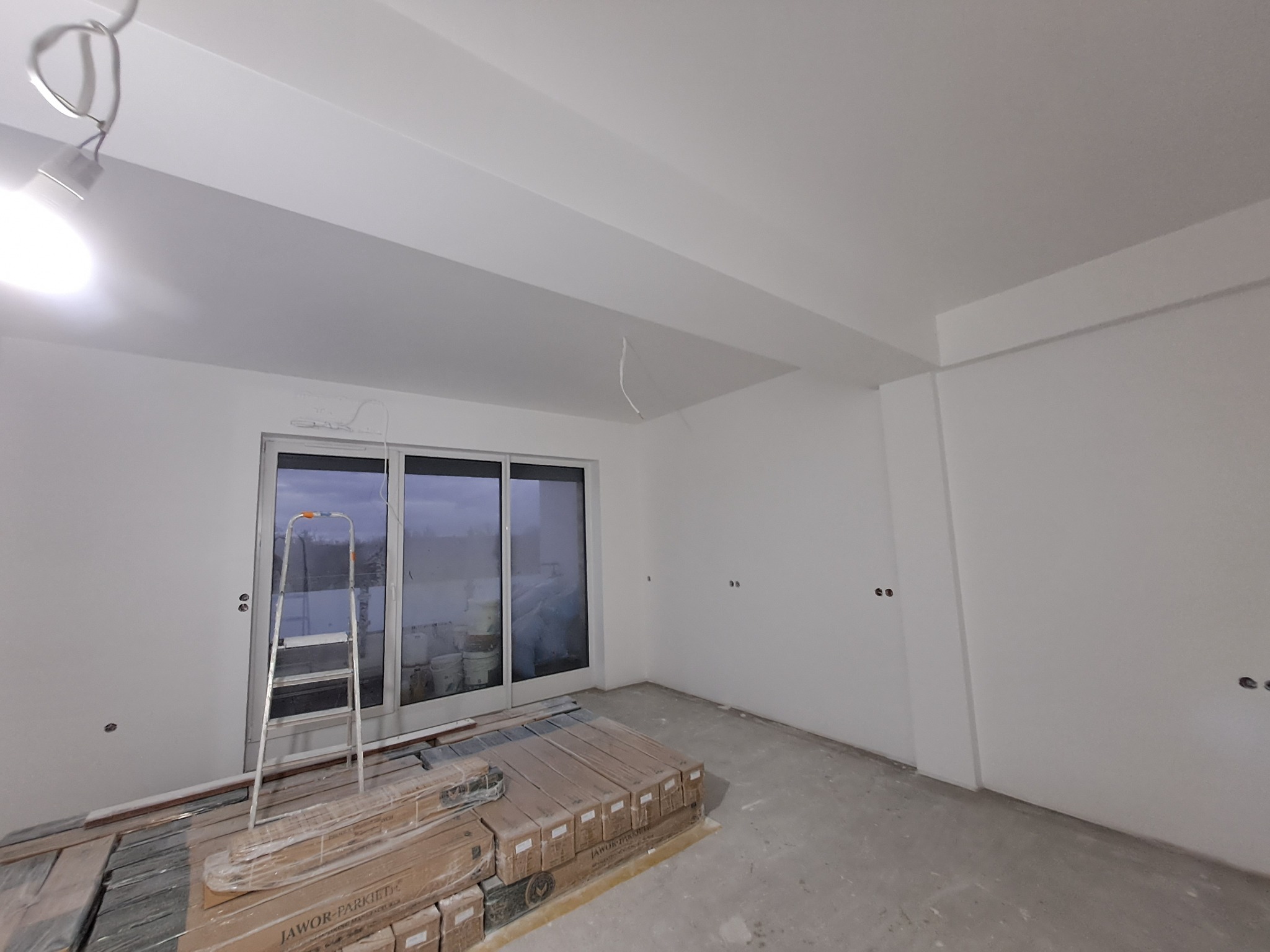 Przygotowanie do układania paneli: stos paczek paneli Jawor-Parkiet na betonowej podłodze, drabina oparta o przesuwne szklane drzwi balkonowe w surowym, białym wnętrzu z widocznymi niedokończonymi...