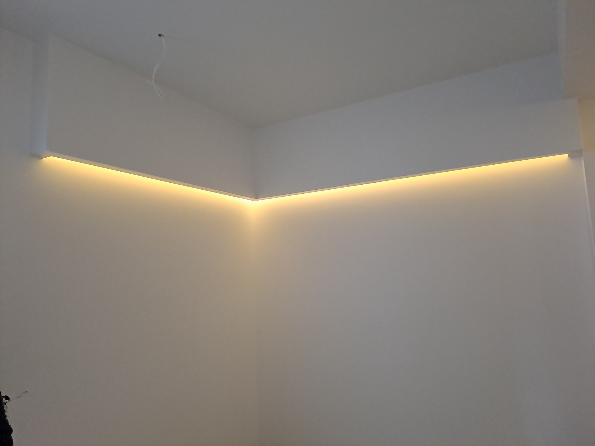 Wnętrze narożne z podwieszanym sufitem i liniowym oświetleniem LED, widoczne przewody elektryczne wystające z sufitu.