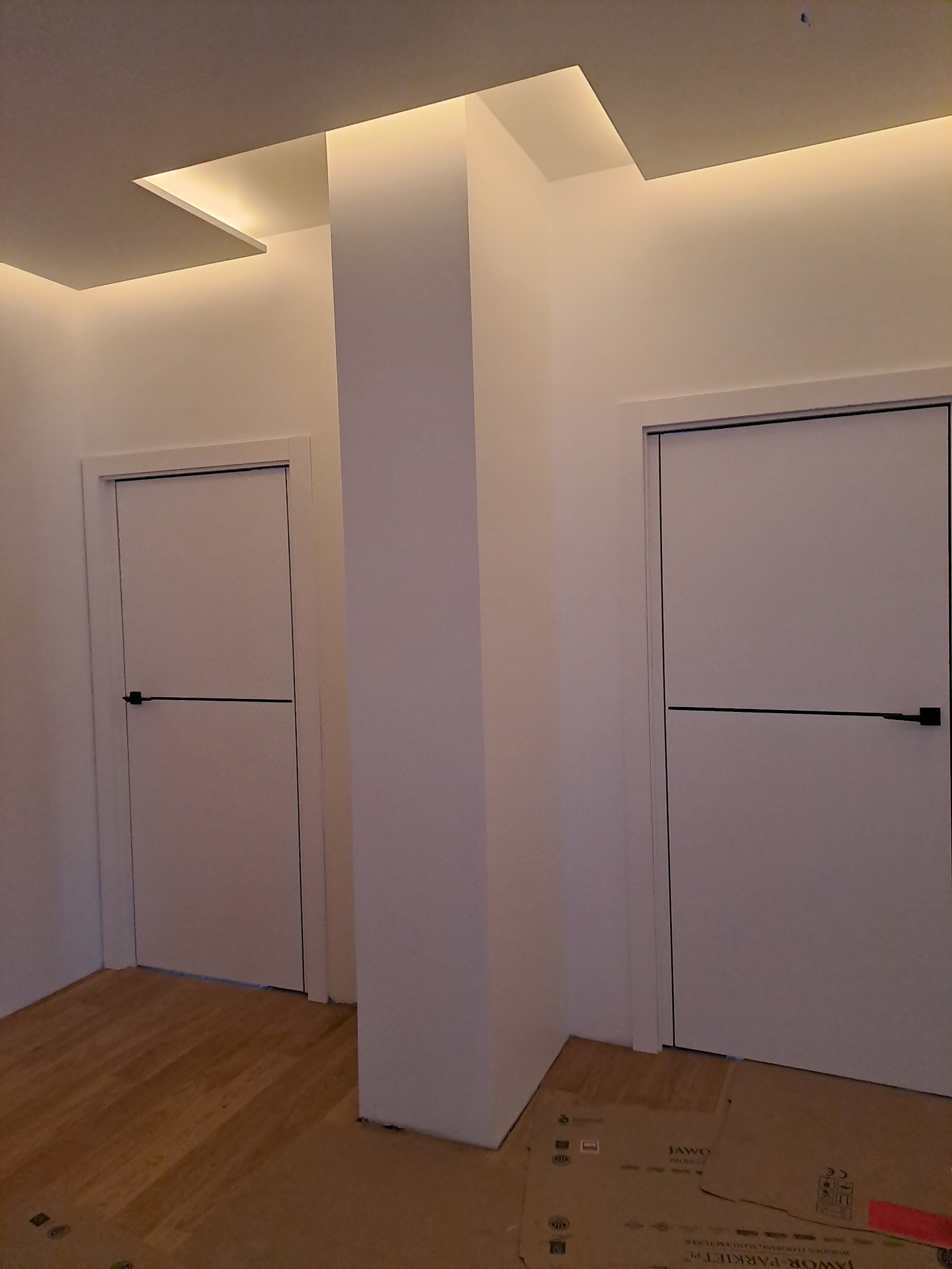 Nowoczesne wnętrze z dwiema białymi drzwiami z czarnymi klamkami, oświetleniem LED w suficie podwieszanym i częściowo ułożonymi panelami podłogowymi, zabezpieczonymi kartonem.