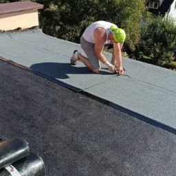 Dekarz w białym podkoszulku i szarych spodenkach, klęczący na dachu pokrytym papą, przycina nożem fragment papy. W tle widoczne drzewa i zabudowania.