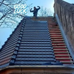 Łukasz Kuczewski - Częściowo pokryty dach z czarną dachówką, widoczne łaty, mężczyzna na szczycie w geście triumfu, Nowa Ruda. Część dachu jeszcze bez pokrycia.